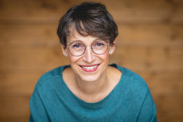 Kopf-und-Schulter-Porträt von Petra Pflugfelder, lächelnden , mit kurz geschnittenem braunen Haar und runden Brillengläsern, frontal vor einem unscharfen Holzhintergrund; sie trägt einen türkisgrünen Pullover und kleine Ohrstecker