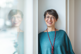 Porträt von Petra Pflugfelder, lächelnde mit kurz geschnittenem dunklem Haar und runder Brille, an einer hellen Wand lehnend; links spiegelt sich ihr Profil im Fenster; sie trägt einen türkisenen Pullover und eine lange rote Perlenkette 